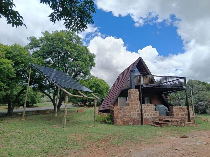 A rustic A-frame house featuring stone masonry, a timber balcony, and a ground-mounted solar panel array set in a natural garden landscape.