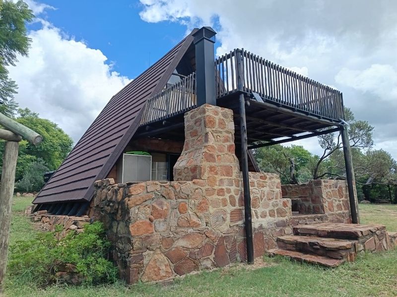 A-frame cabin featuring a stone chimney, elevated timber balcony, and rustic stone masonry exterior set within a natural garden environment.