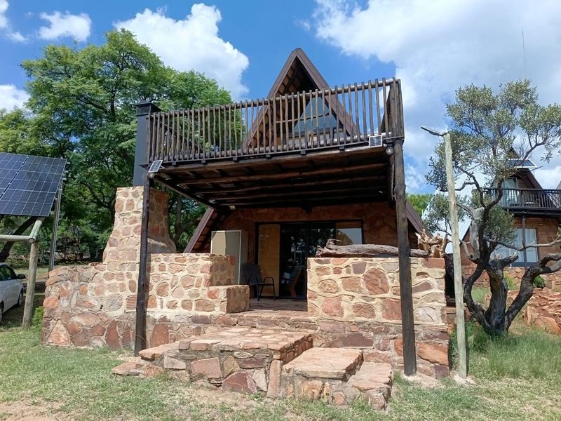 A rustic A-frame house featuring stone masonry walls, a raised timber balcony, and integrated solar panels set within a natural garden environment.