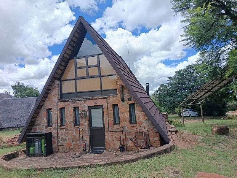 A unique A-frame house featuring stone cladding, a large triangular window, and a stone patio area set in a natural landscape.