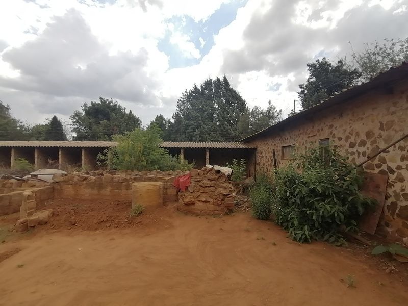 Rear exterior view of a property featuring rustic stone masonry walls and a corrugated roof structure with surrounding unpaved grounds.