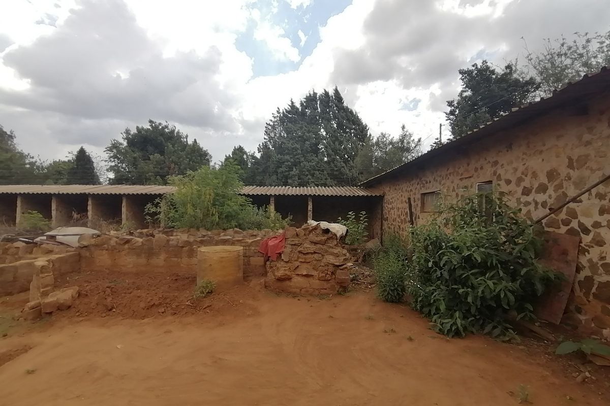 Rear exterior view of a property featuring rustic stone masonry walls and a corrugated roof structure with surrounding unpaved grounds.
