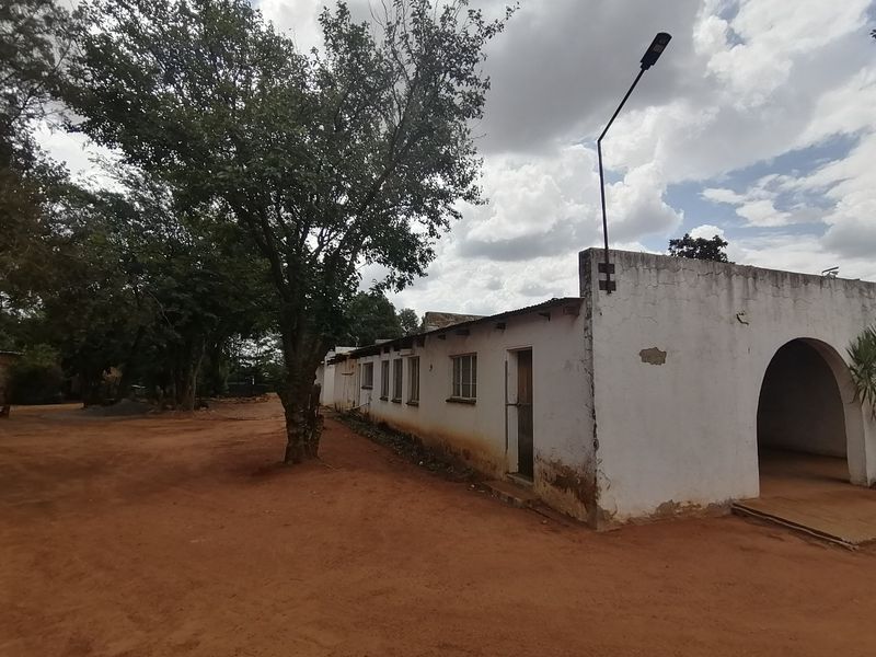 Exterior view of a single-story house featuring an arched entrance, weathered white plaster walls, and a metal roof set on a dirt lot.