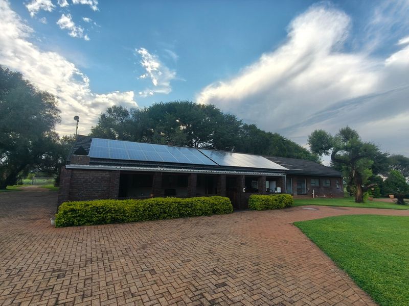 A well-maintained farm property featuring a brick exterior, a covered patio area, and a roof equipped with solar panels, set on a paved driveway.