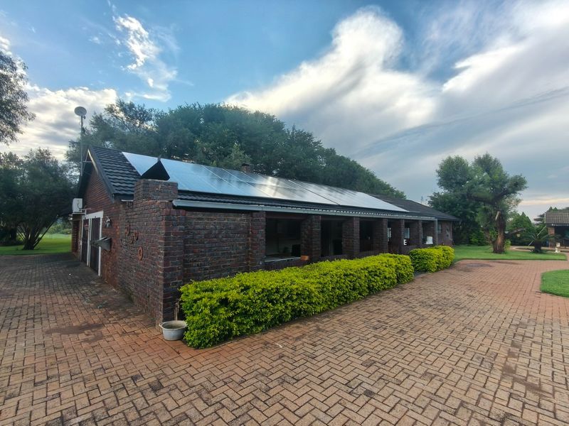A rear view of a brick farm building featuring a large solar panel array on the roof and an expansive paved driveway area.