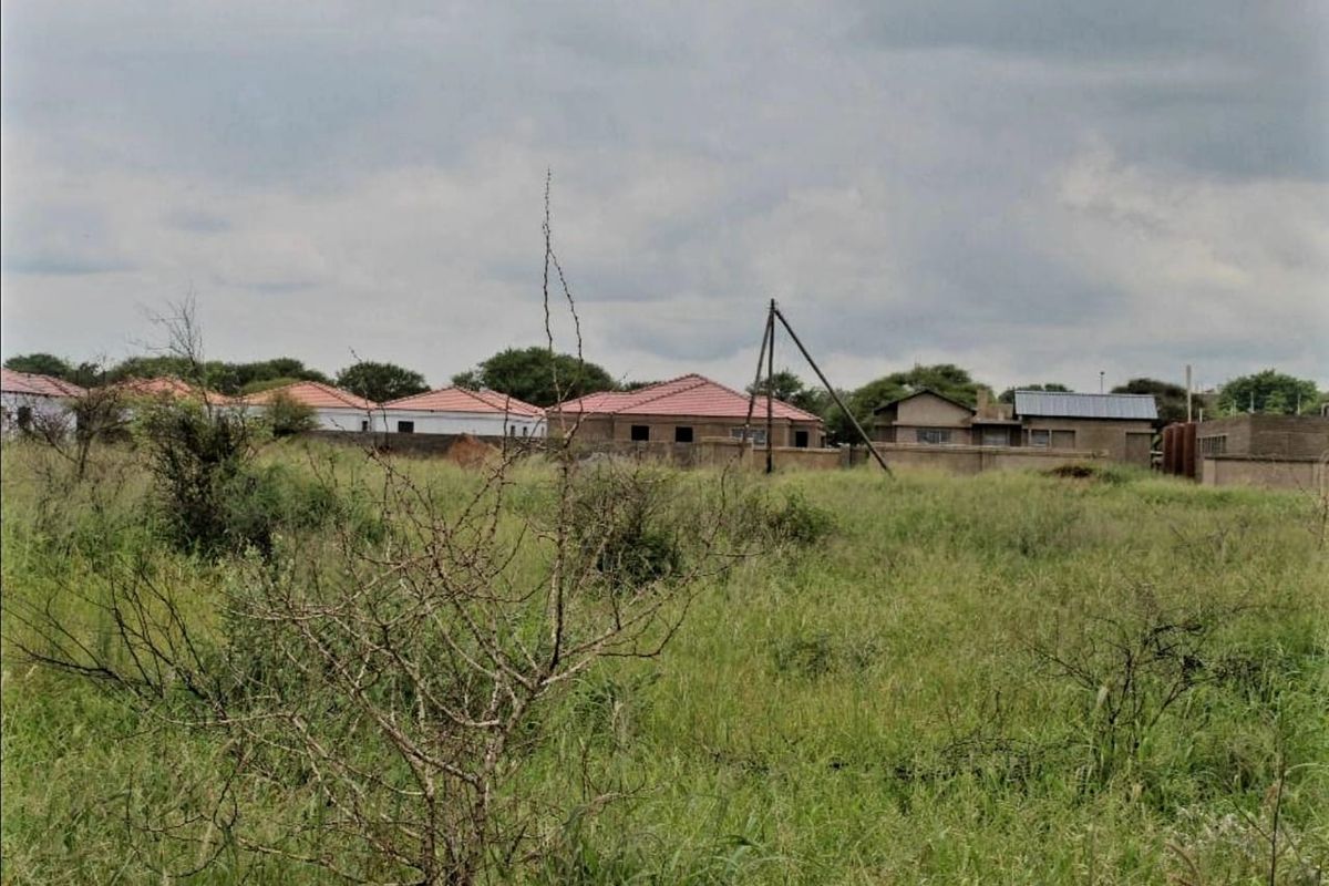 A view of a vacant residential plot featuring overgrown grass with neighboring houses and a perimeter wall visible in the background.