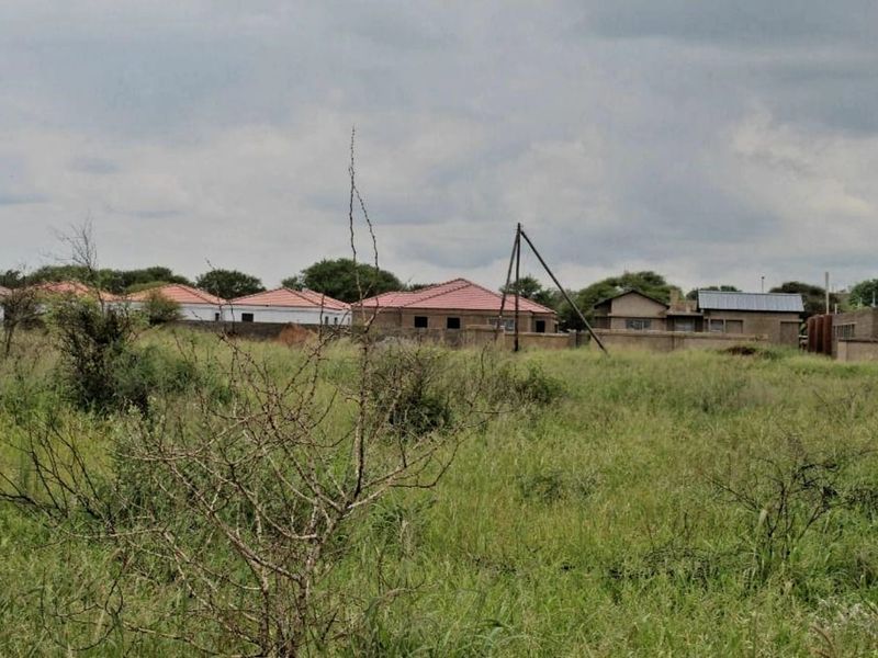 A view of a vacant residential plot featuring overgrown grass with neighboring houses and a perimeter wall visible in the background.