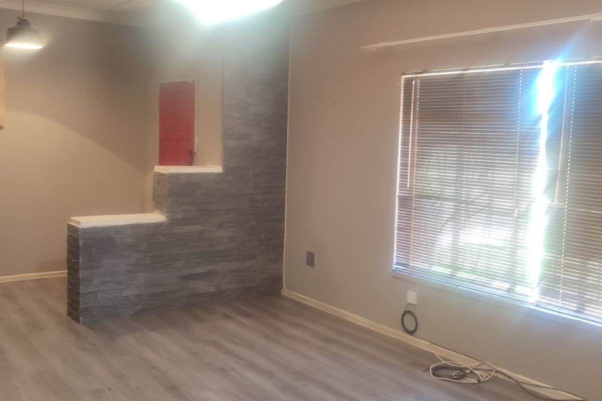 A spacious living area featuring modern laminate flooring, a decorative stone-cladding feature wall, and large windows fitted with venetian blinds.