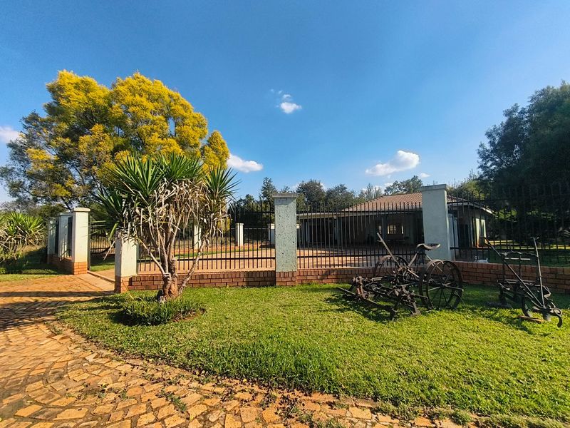 Front view of the farm property featuring a paved driveway, landscaped garden, and secure perimeter fencing with brick and metal construction.
