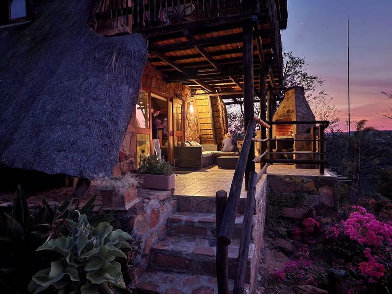 A rustic covered patio featuring stone masonry, a built-in fireplace, and a thatched roof, offering a charming outdoor living space with sunset views.