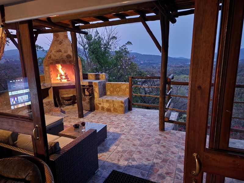 A covered outdoor patio featuring a stone fireplace, tiled flooring, and timber structural elements, offering scenic elevated views of the surrounding landscape.