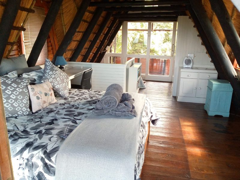 A rustic loft-style bedroom featuring exposed timber rafters, a traditional thatch roof, timber flooring, and direct access to an outdoor balcony area.