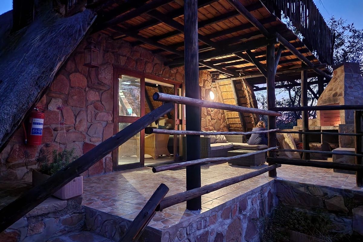 A rustic covered patio featuring stone walls, timber support beams, tiled flooring, and a built-in fireplace, providing a cozy outdoor entertainment space.