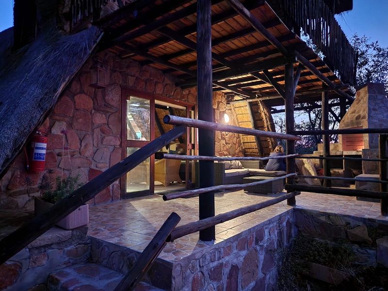 A rustic covered patio featuring stone walls, timber support beams, tiled flooring, and a built-in fireplace, providing a cozy outdoor entertainment space.