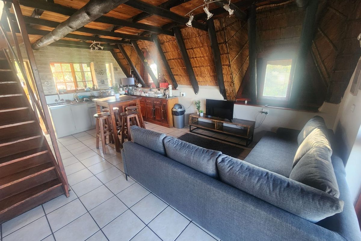 Open-plan living area featuring exposed timber rafters, a thatched ceiling, tiled flooring, and an integrated kitchen space with a wooden staircase.