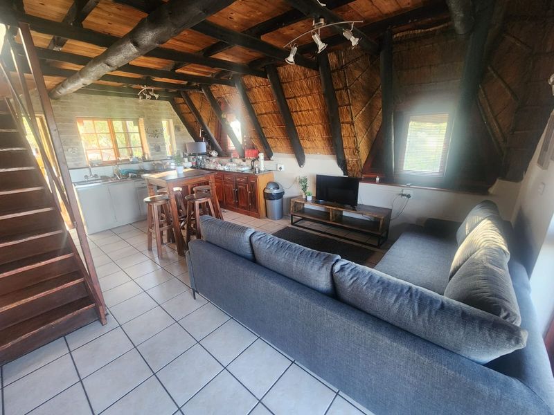 Open-plan living area featuring exposed timber rafters, a thatched ceiling, tiled flooring, and an integrated kitchen space with a wooden staircase.