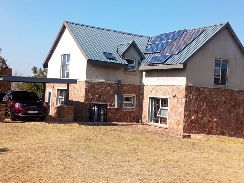 A two-story house featuring stone cladding, a metal roof with integrated solar panels, and an attached carport, set in a rural landscape.