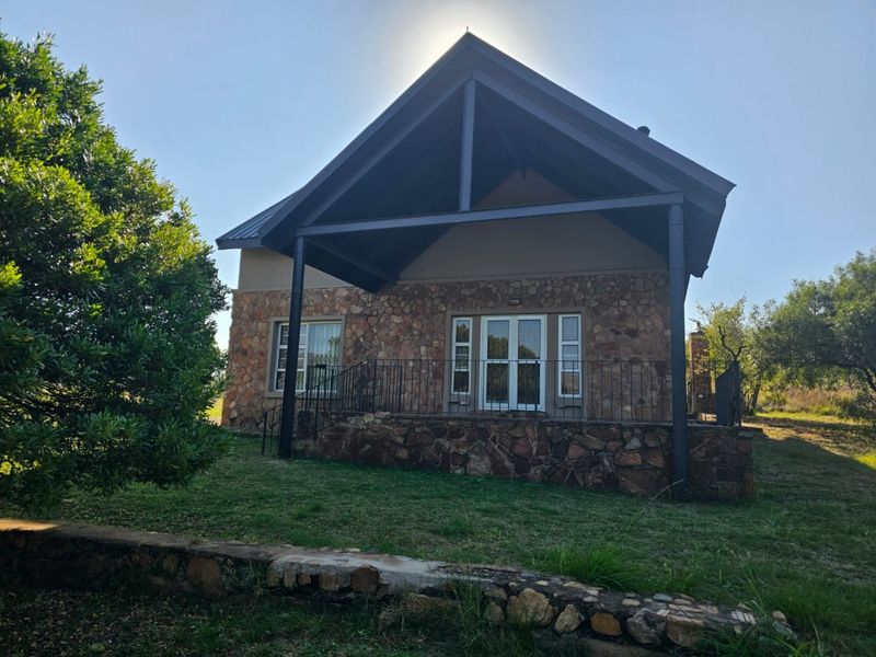 A charming stone-clad house featuring a covered patio, metal roof, and wrought iron railings, set within a natural garden landscape.