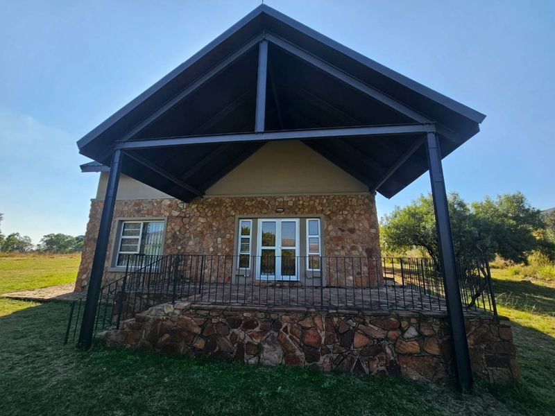 A rear view of the house featuring a raised stone patio, covered by a high-pitched metal roof supported by steel beams.