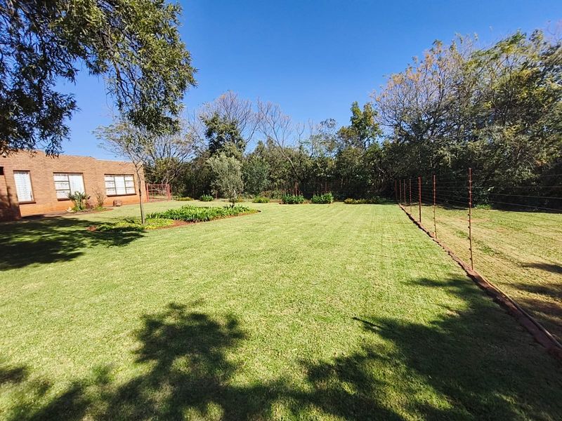 A well-maintained, expansive lawn area featuring established garden beds, brick building exterior, and perimeter electric fencing.