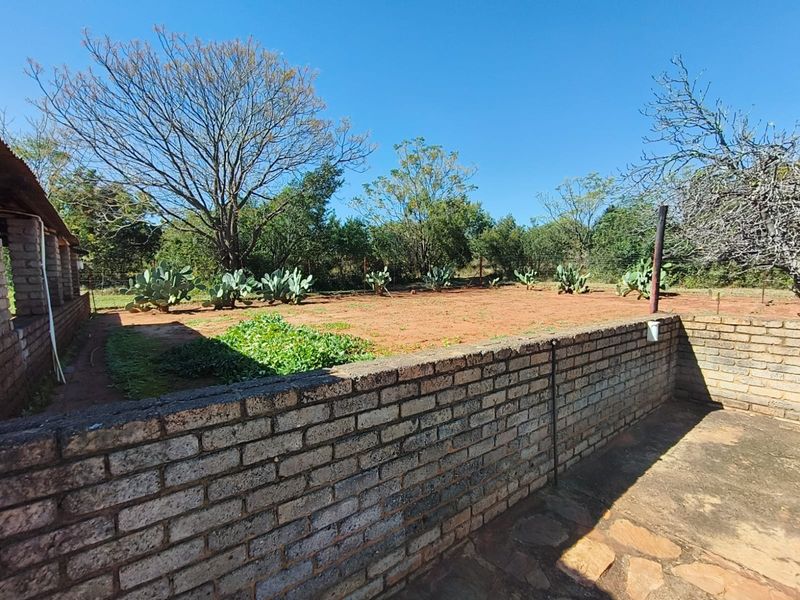A rear exterior view of the farm property featuring a brick boundary wall, stone paved area, and open garden space with mature trees.