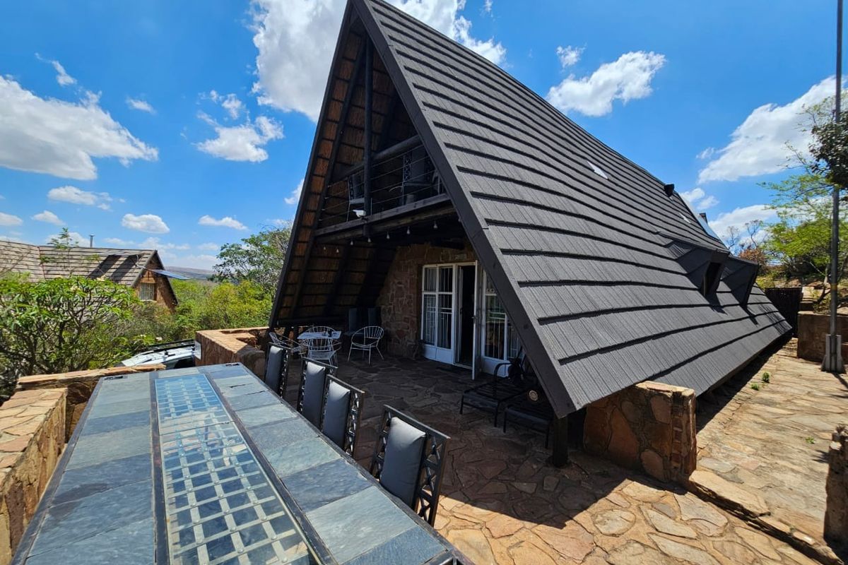 A rear view of an A-frame house featuring stone cladding, a large tiled roof, a balcony, and a spacious stone-paved patio area.