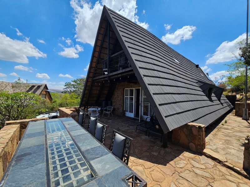 A rear view of an A-frame house featuring stone cladding, a large tiled roof, a balcony, and a spacious stone-paved patio area.