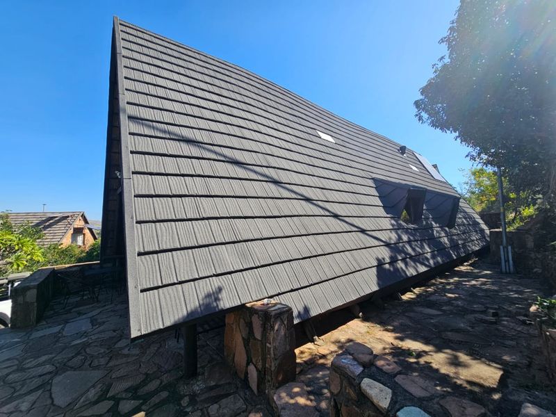 A unique A-frame house exterior featuring a steep tiled roof, stone masonry foundation, and integrated dormer windows set against a stone-paved patio area.