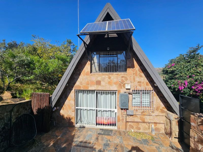 A-frame house exterior featuring stone cladding, a tiled patio, security bars on windows, and roof-mounted solar panels.