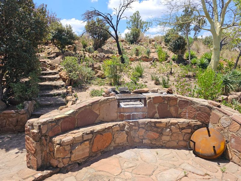 Outdoor patio area featuring a stone-built seating wall, integrated braai, and stone stairs leading up a landscaped hillside.