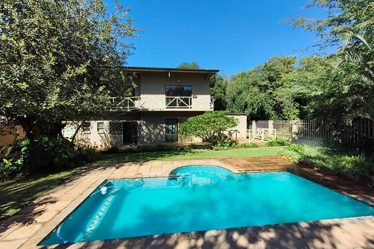 Rear view of a two-story house featuring a swimming pool, paved deck, and a balcony with wooden railings, set within a landscaped garden.