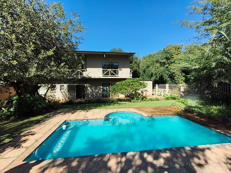 Rear view of a two-story house featuring a swimming pool, paved deck, and a balcony with wooden railings, set within a landscaped garden.