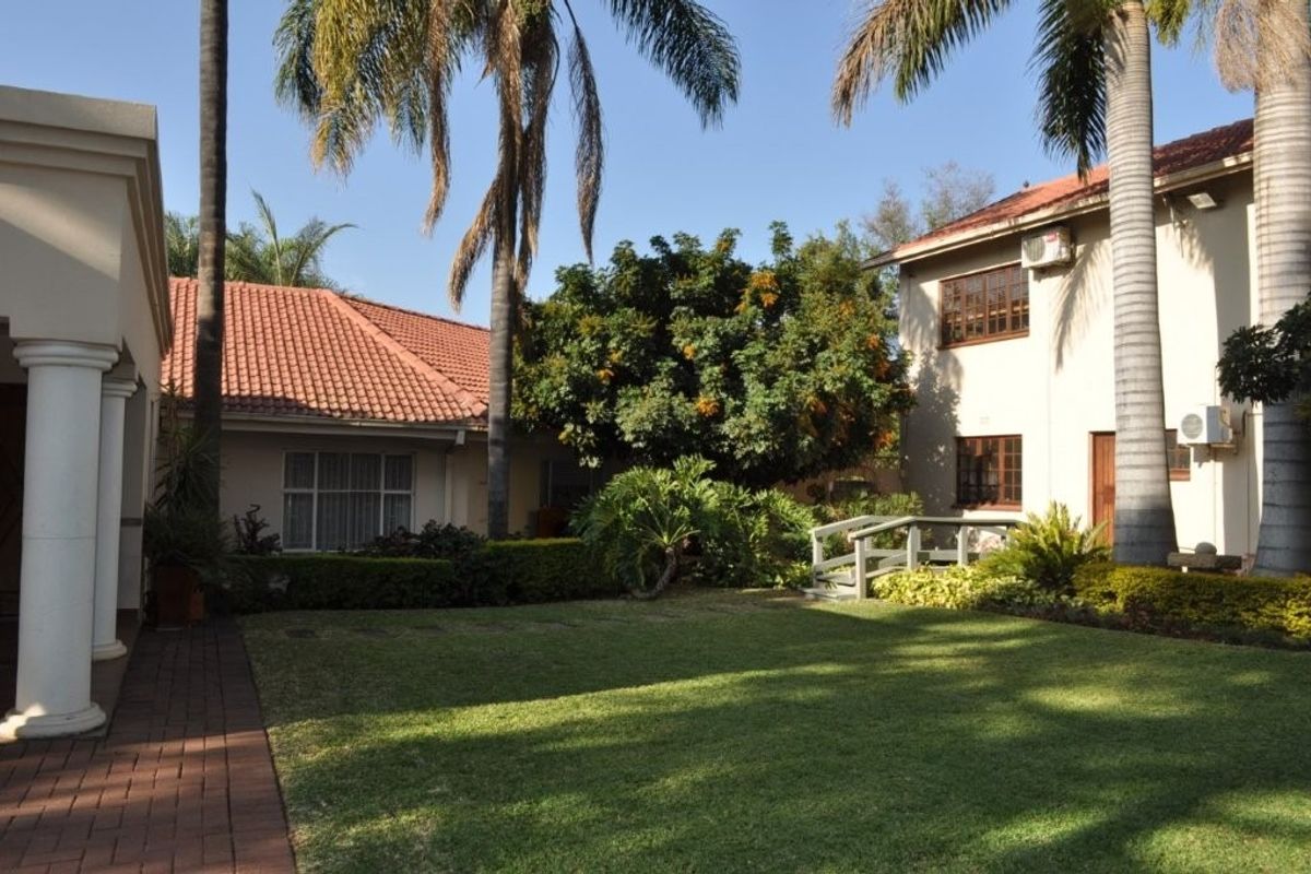 A well-maintained rear garden area featuring a lush lawn, mature palm trees, a small pedestrian bridge, and terracotta-tiled roof structures.