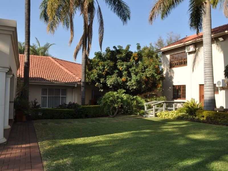 A well-maintained rear garden area featuring a lush lawn, mature palm trees, a small pedestrian bridge, and terracotta-tiled roof structures.