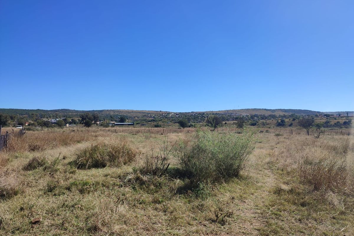 A wide-angle view of a vacant residential plot featuring natural dry grass, sparse vegetation, and distant hillside landscape views.