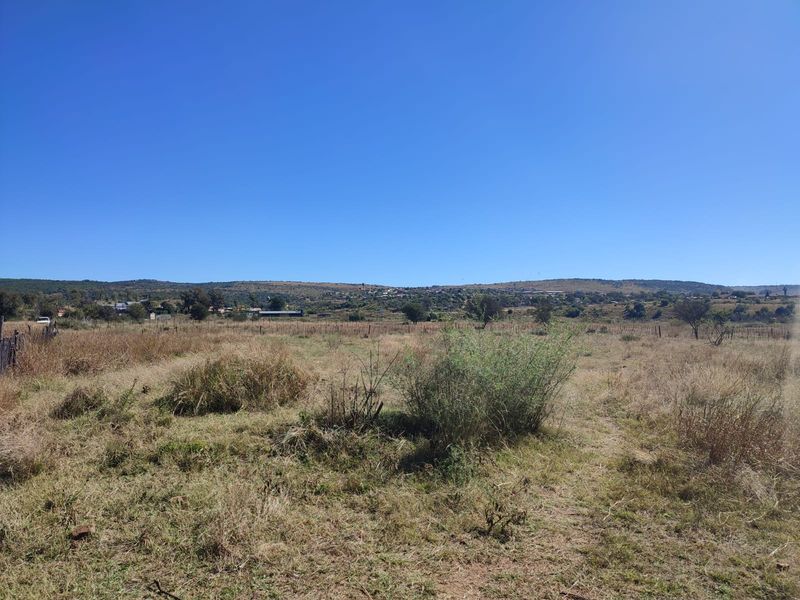 A wide-angle view of a vacant residential plot featuring natural dry grass, sparse vegetation, and distant hillside landscape views.