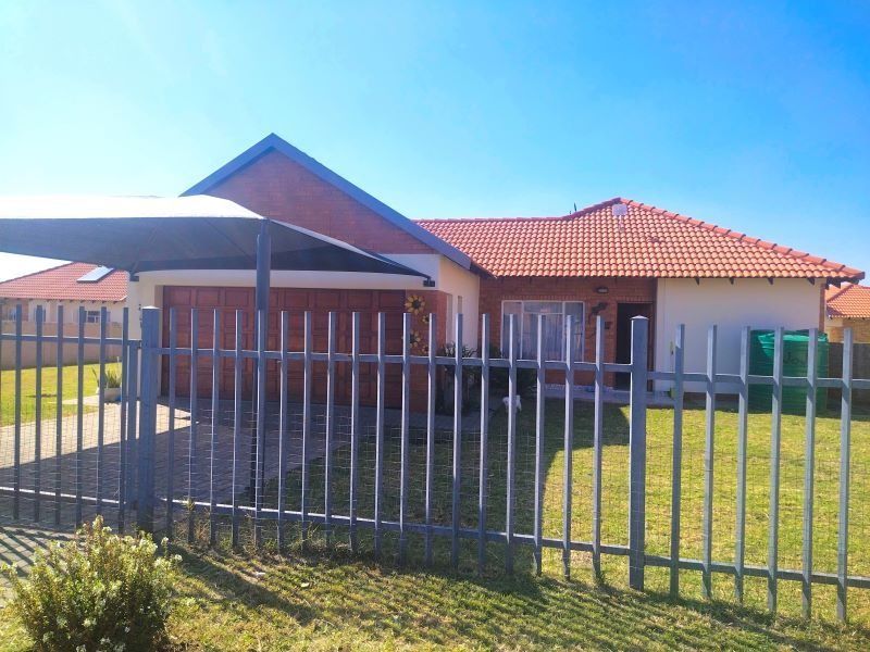 Front view of a single-story brick house featuring a tiled roof, paved driveway with a carport, and secure steel perimeter fencing.