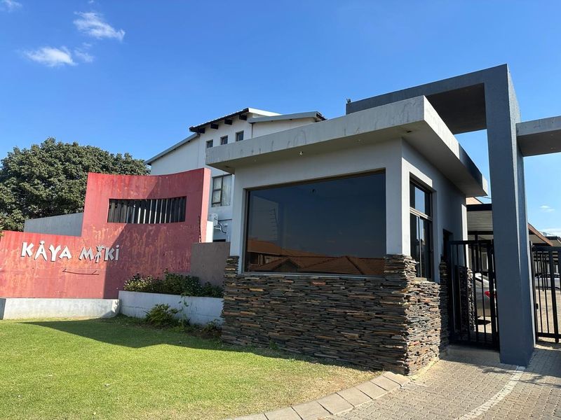 Front entrance view of the Kaya Maki complex featuring a security guard house with stone cladding, paved driveway, and a prominent boundary wall.