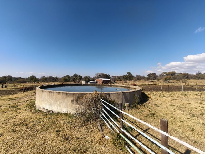 A large circular concrete water reservoir situated in an open farm field, featuring metal perimeter fencing and clear blue sky.