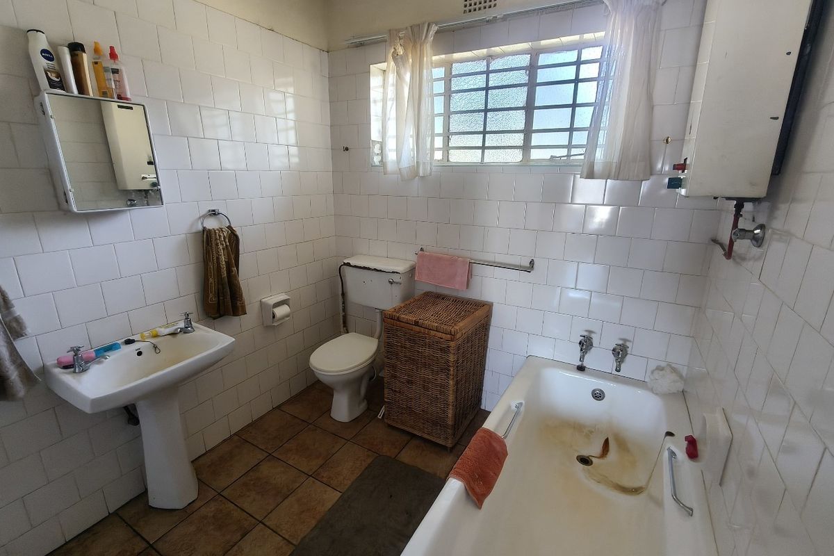 A dated bathroom featuring a pedestal basin, toilet, and bathtub with full-height wall tiling and a window with security bars.