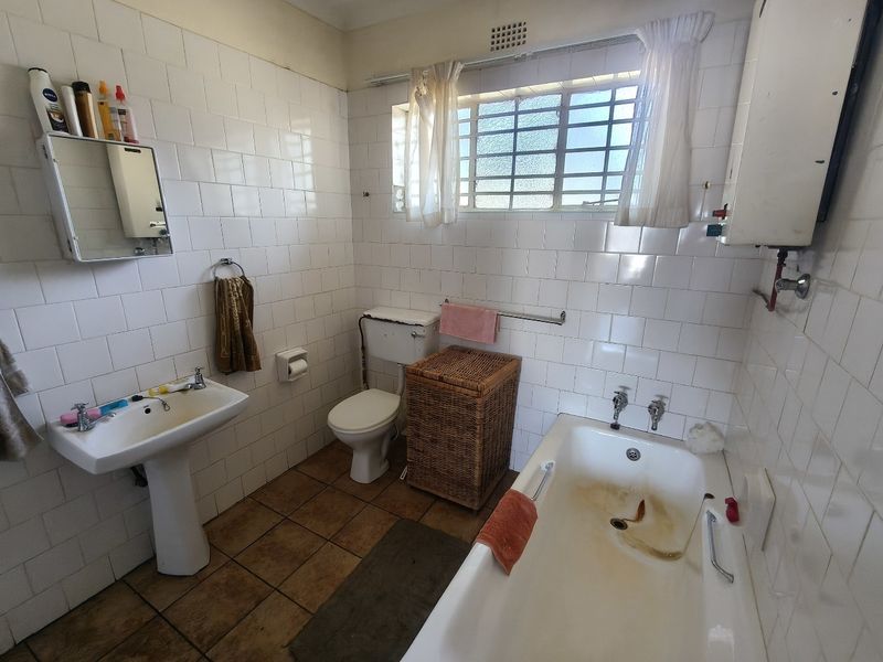 A dated bathroom featuring a pedestal basin, toilet, and bathtub with full-height wall tiling and a window with security bars.