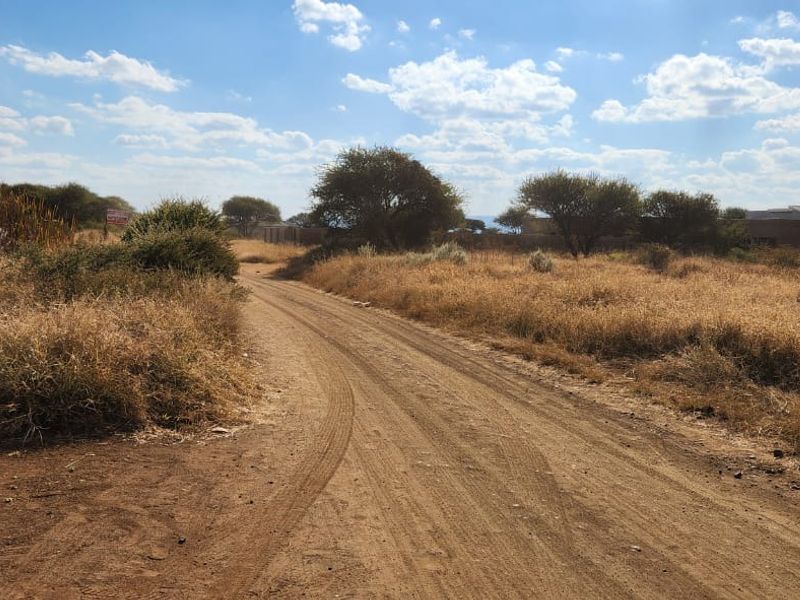 A view of an unpaved access road leading through a vacant residential plot with native vegetation and surrounding landscape.