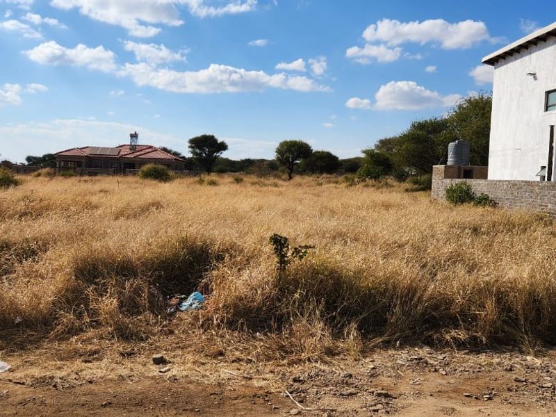 A view of a vacant residential plot featuring overgrown grass, a brick boundary wall, and an adjacent building with a water tank.