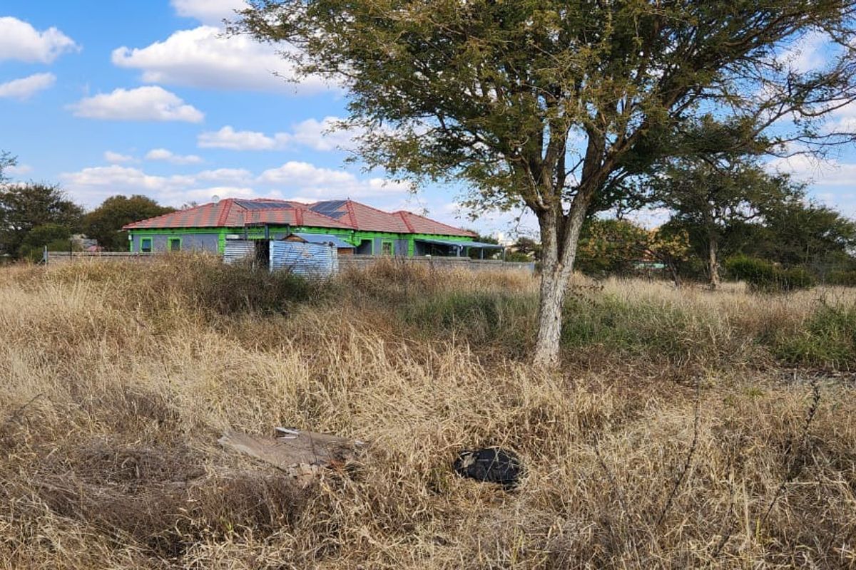 A view of a vacant residential plot featuring mature trees and surrounding fencing, with neighboring properties visible in the background.