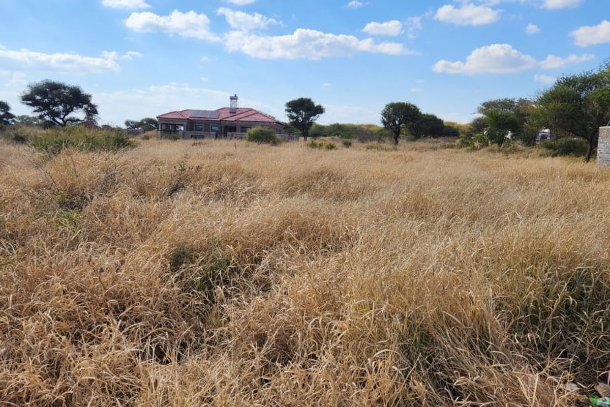 A view of a vacant residential plot featuring tall dry grass, native trees, and a small brick structure with a water storage tank.