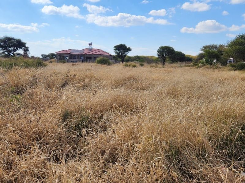 A view of a vacant residential plot featuring tall dry grass, native trees, and a small brick structure with a water storage tank.
