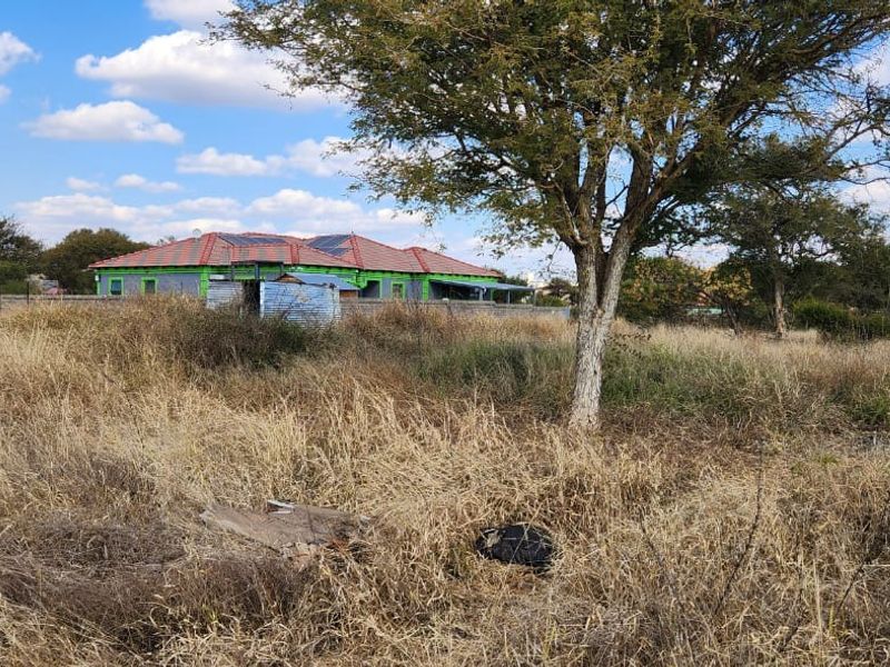 A view of a vacant residential plot featuring mature trees and surrounding fencing, with neighboring properties visible in the background.