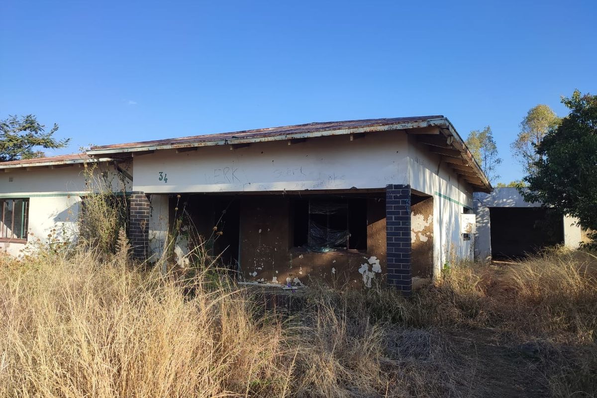 Front elevation of a single-story house in poor condition, featuring a corrugated metal roof, brick pillars, and significant exterior wall damage.