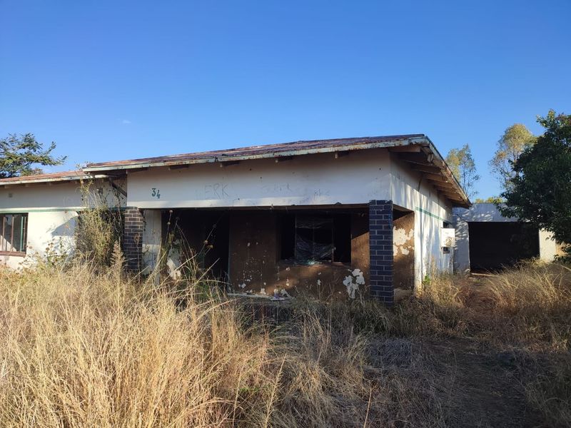 Front elevation of a single-story house in poor condition, featuring a corrugated metal roof, brick pillars, and significant exterior wall damage.