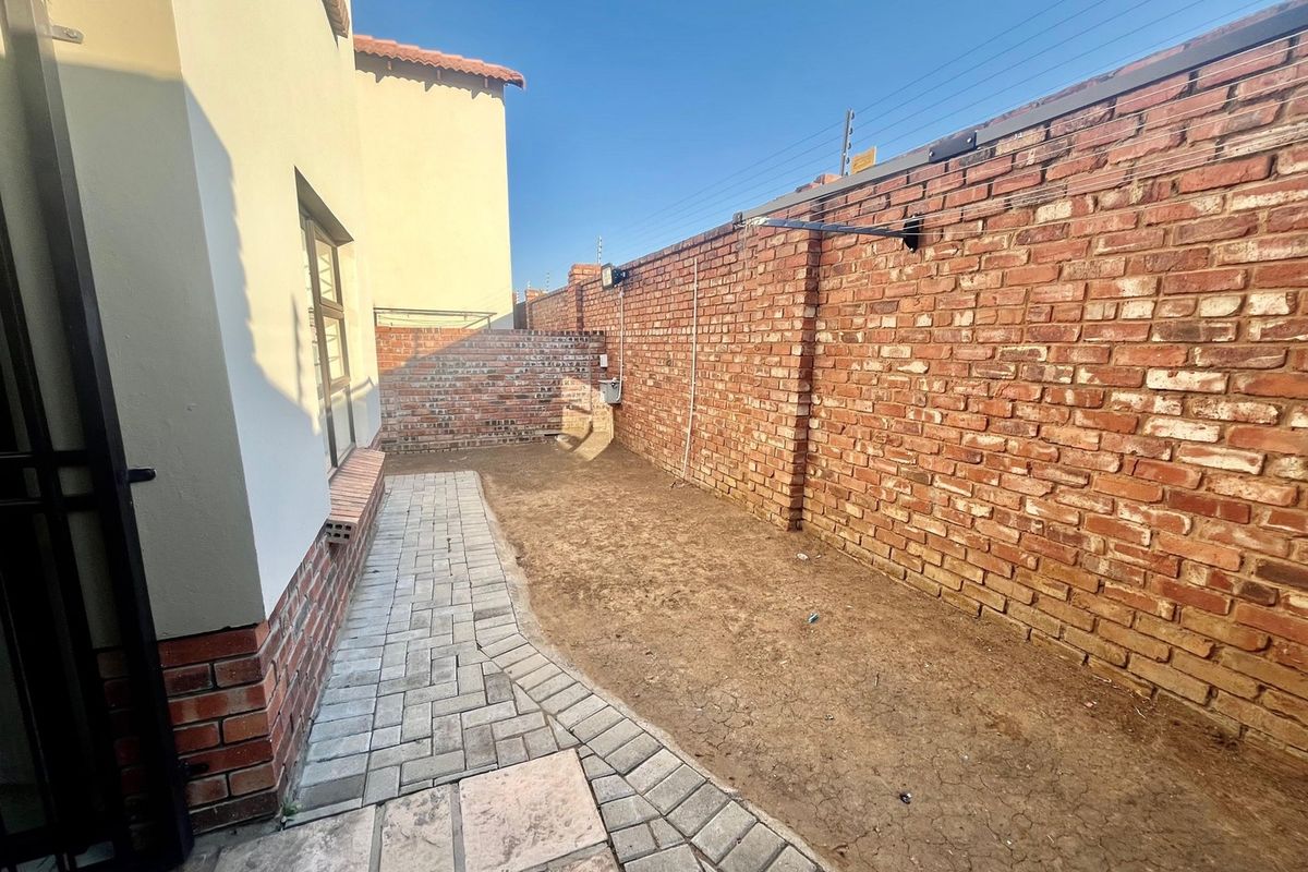 A rear exterior area featuring a paved walkway, brick boundary walls with electric fencing, and an unlandscaped soil garden space.